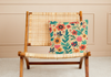 Faux suede pillow with mid century boho inspired floral reds, greens and pinks design on a rattan chair